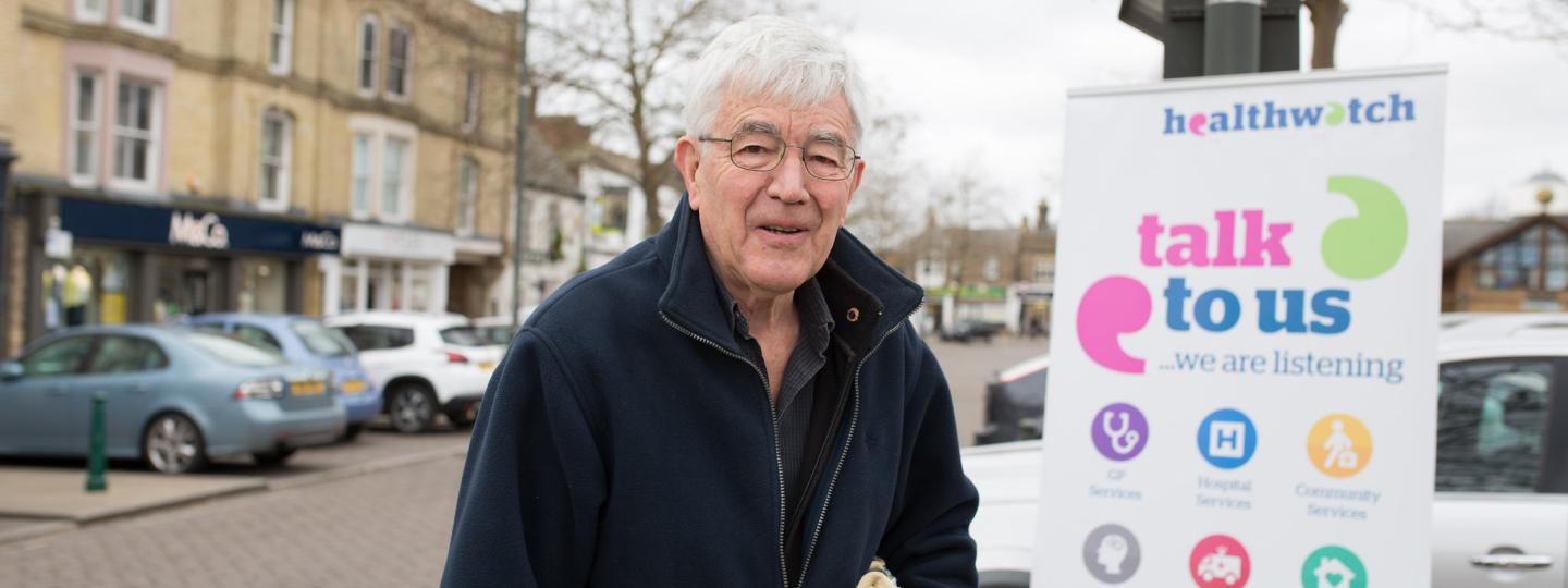 Elderly man standing in front of a Healthwatch banner saying "Talk to us"